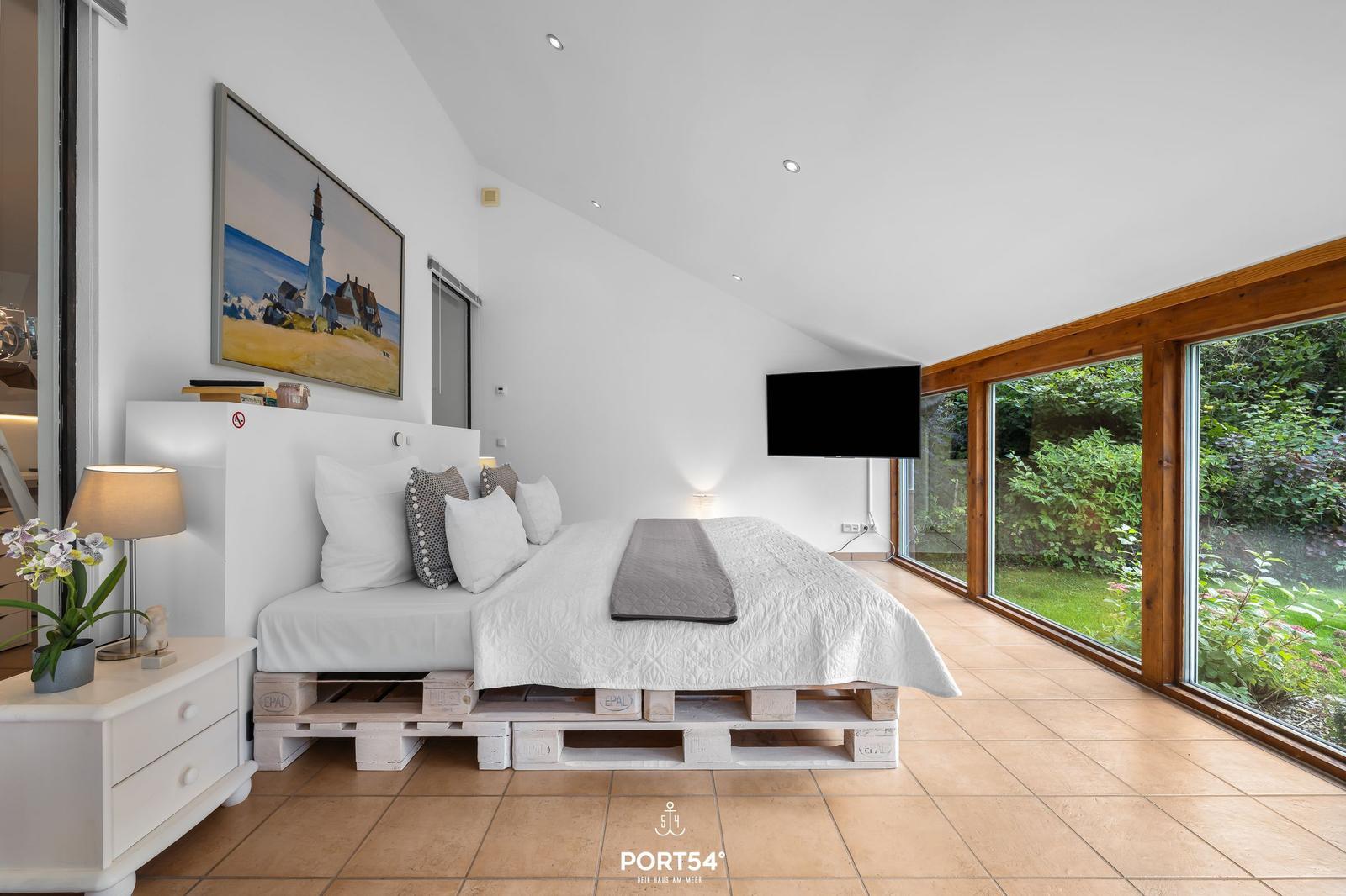 Schlafzimmer mit Bett auf Paletten, Fernseher und großen Fenstern zum Garten.