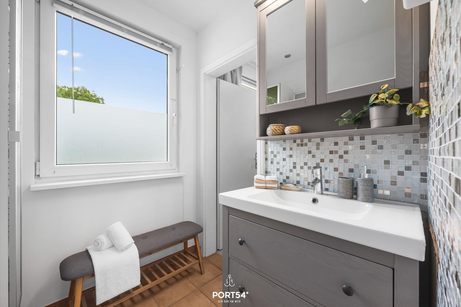 Badezimmer mit Waschtisch, Spiegelschrank und Fenster mit Blick auf Außenbereich.