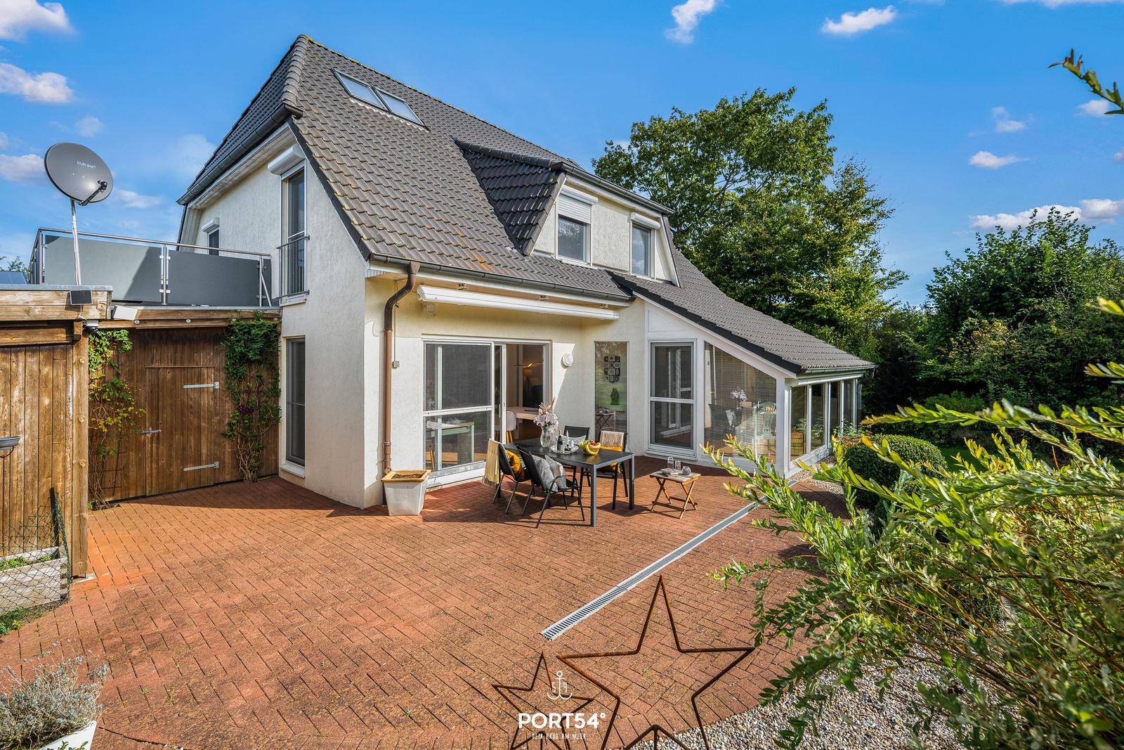 Haus mit Terrasse, Garten und Sicht auf Bäume unter blauem Himmel.