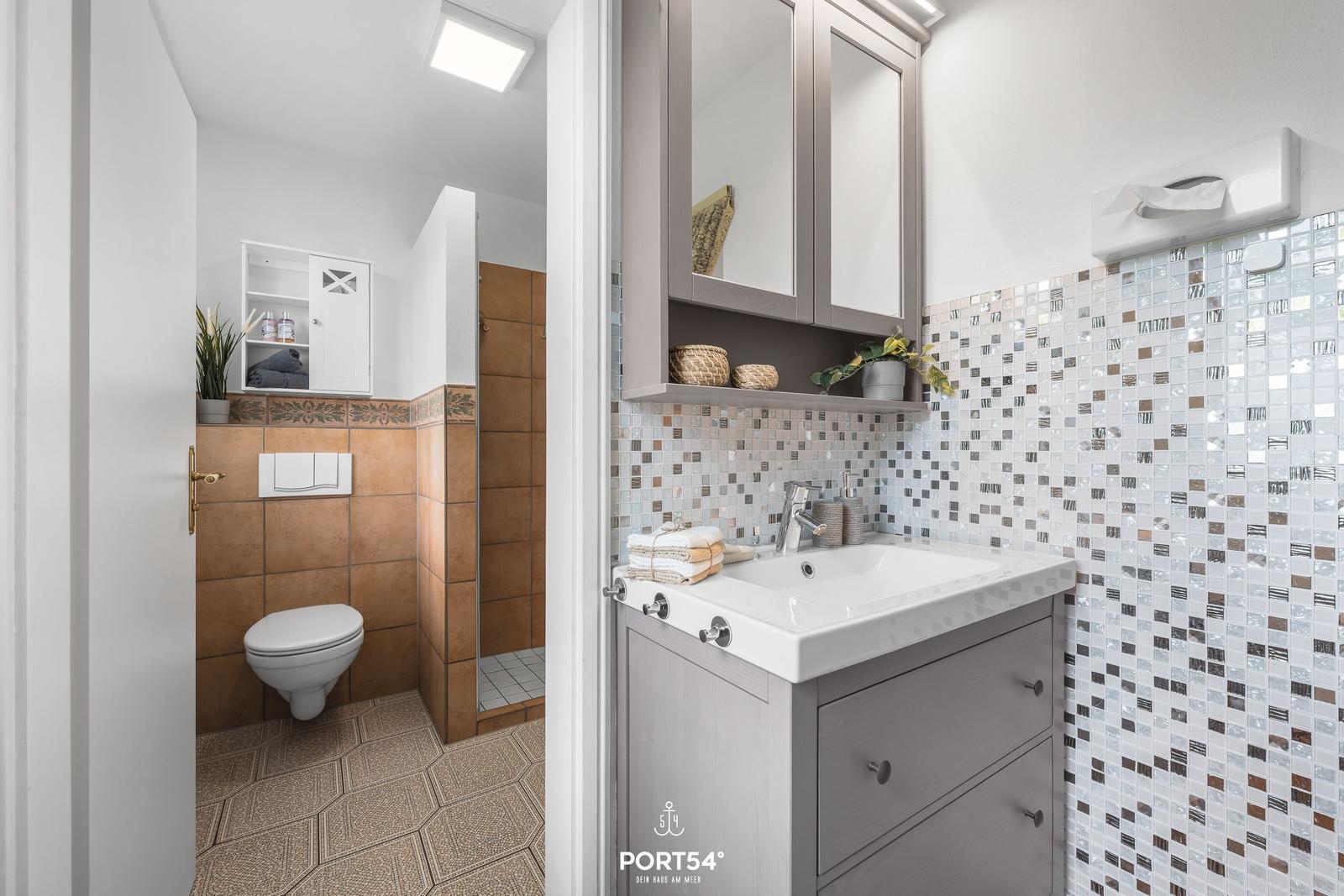 Bad mit Waschbecken, Spiegelschrank und Dusche. Fliesenwand und Toilette sind sichtbar.