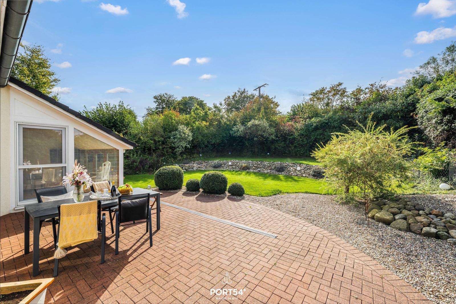 Terrasse mit Tisch und Stühlen, grüner Garten und Steinmauer im Hintergrund.