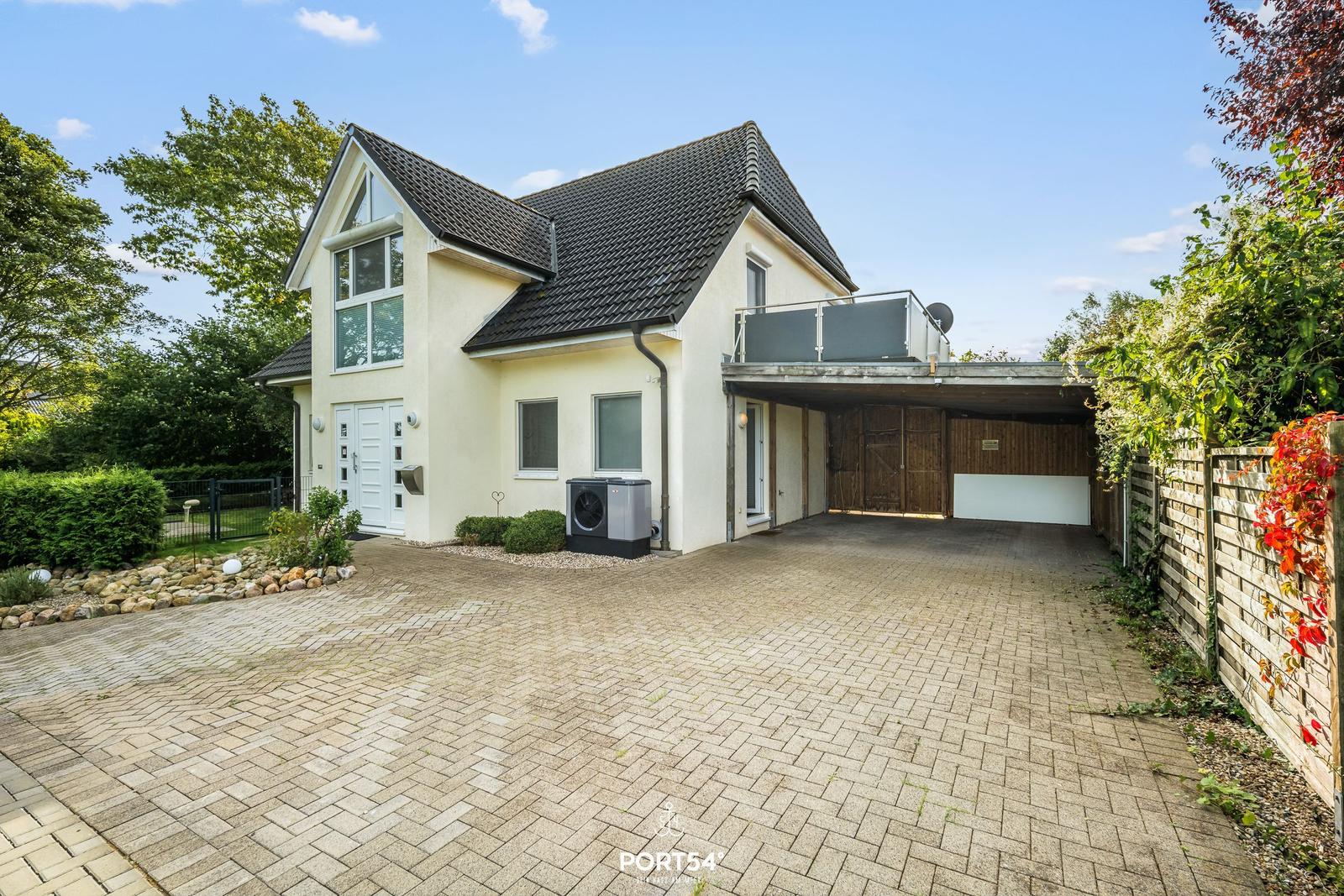 Haus mit Garage, Balkon und gepflastertem Hof. Umgeben von Grün und einem Holzzaun.