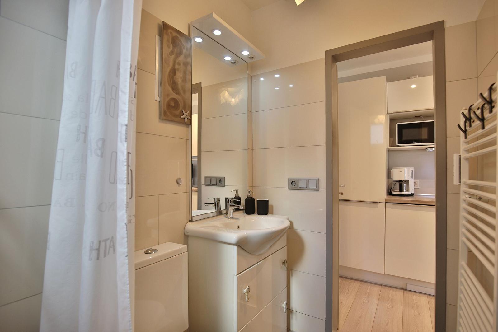 Badezimmer mit Waschbecken, Spiegel und Dusche. Küche mit Mikrowelle und Kaffeemaschine sichtbar.