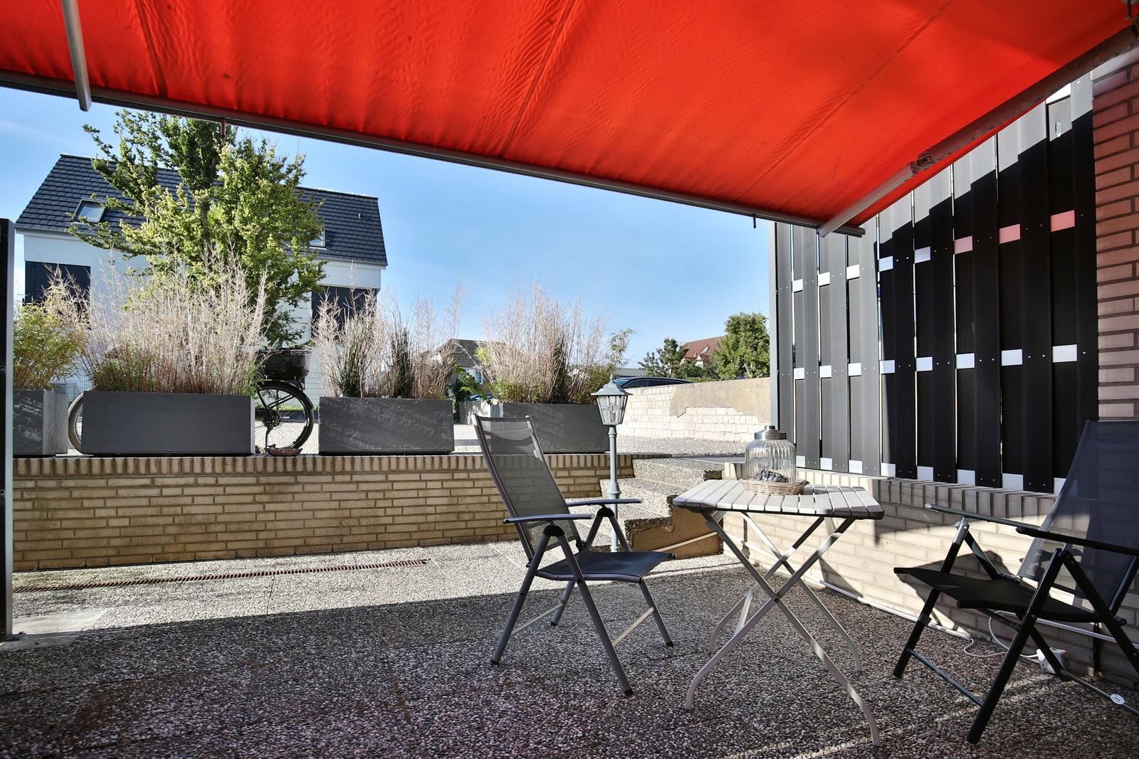 Terrasse mit rotem Sonnensegel, Tisch und zwei Stühlen. Hintergrund: Garten und Nachbarhaus.