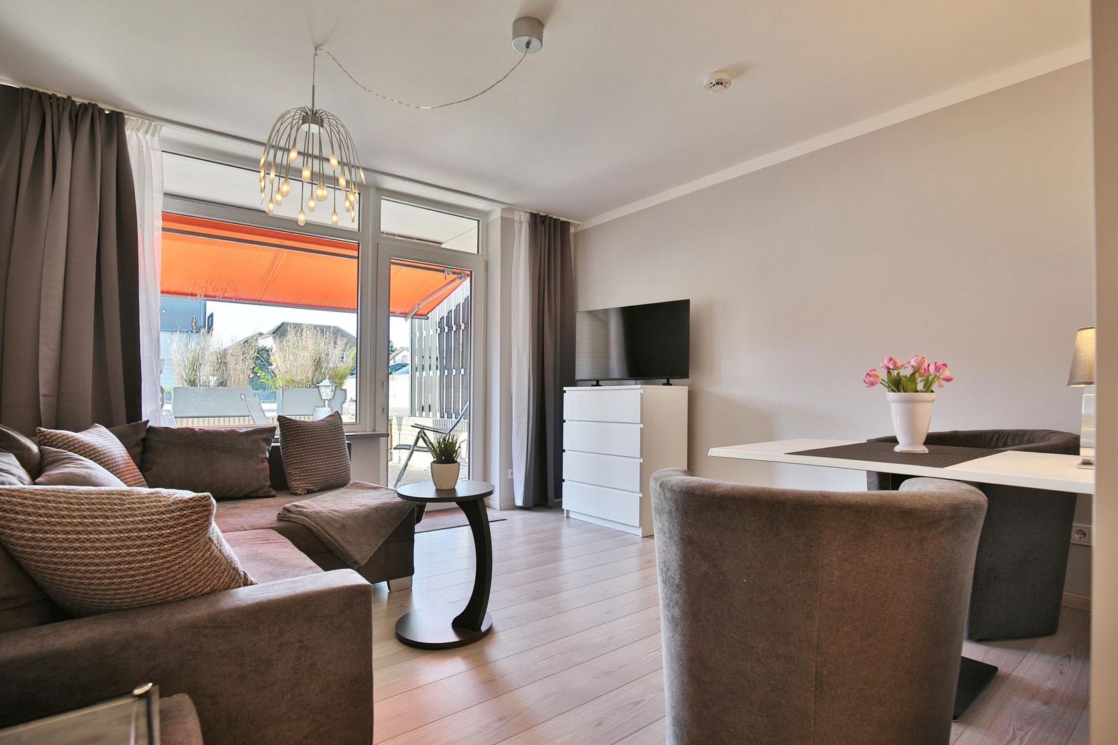 Wohnzimmer mit Sofa, Esstisch und TV. Durchgang zur Terrasse mit orangefarbenem Sonnenschutz.