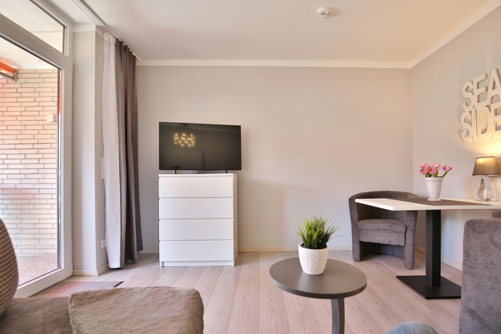 Wohnzimmer mit TV, Schreibtisch und Sitzgelegenheiten. Fenster zur Terrasse.