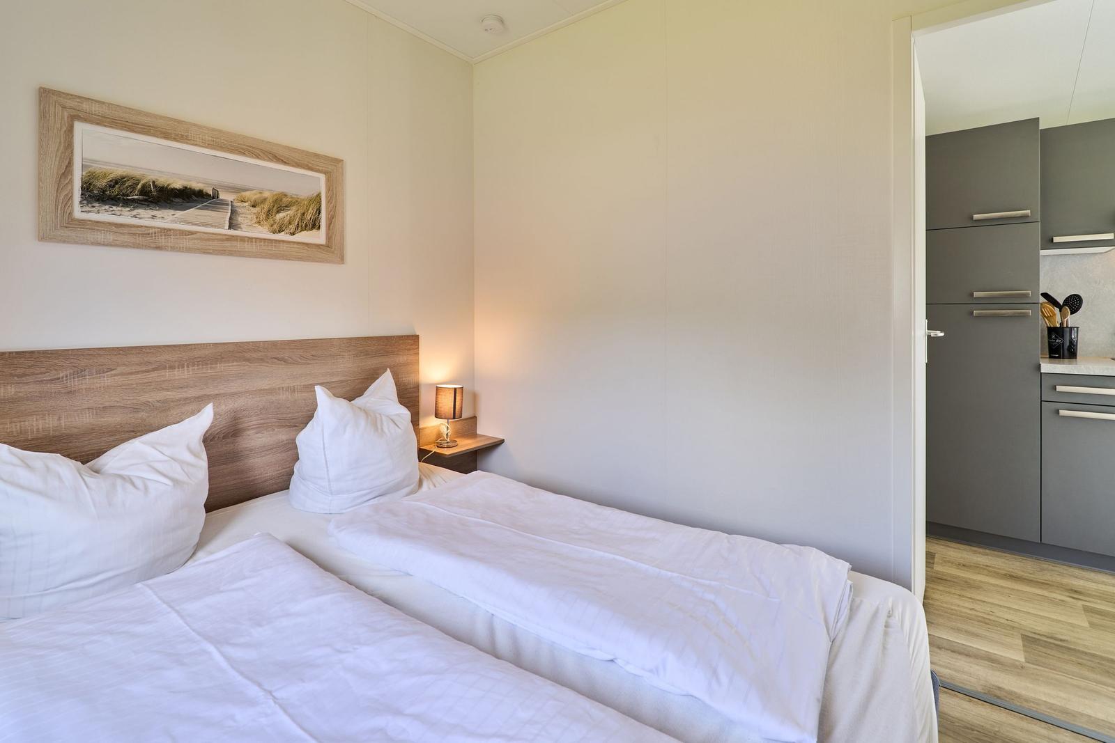 Schlafzimmer mit Doppelbett, Nachttisch und Bild an der Wand. Küche ist im Hintergrund sichtbar.