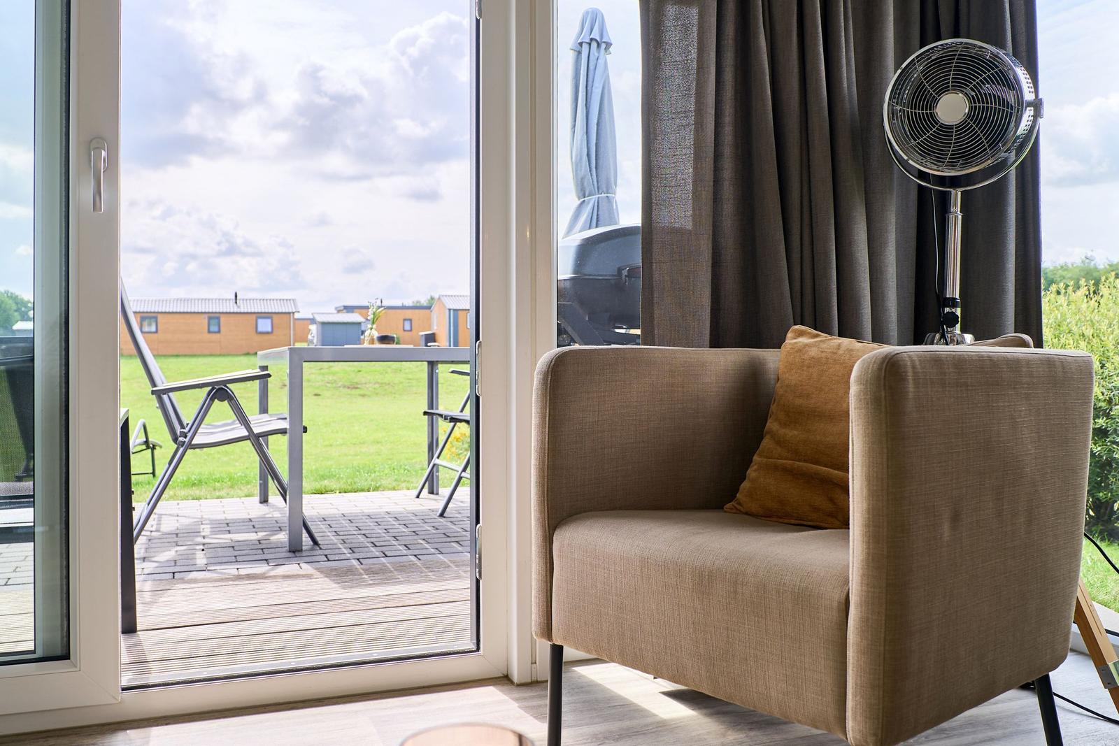 Sitzgelegenheit mit Kissen und Ständerventilator vor Glasfront mit Blick auf Terrasse und Grünfläche.