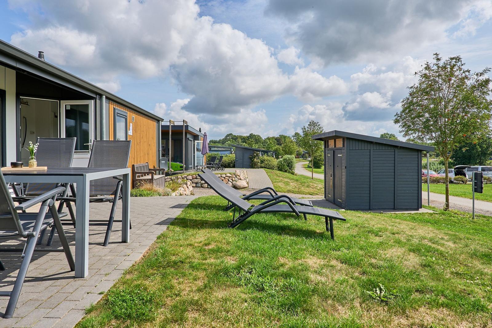 Terrasse mit Tisch und Stühlen, Liegestühle auf Rasen, Gartenhaus und Ferienhaus im Hintergrund.