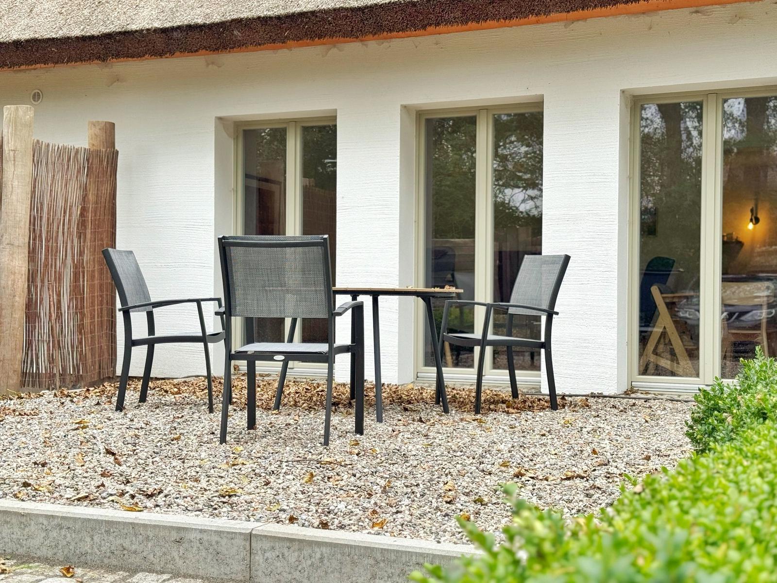 Terrasse mit Tisch und drei Stühlen vor einem Haus mit Schiebetüren.