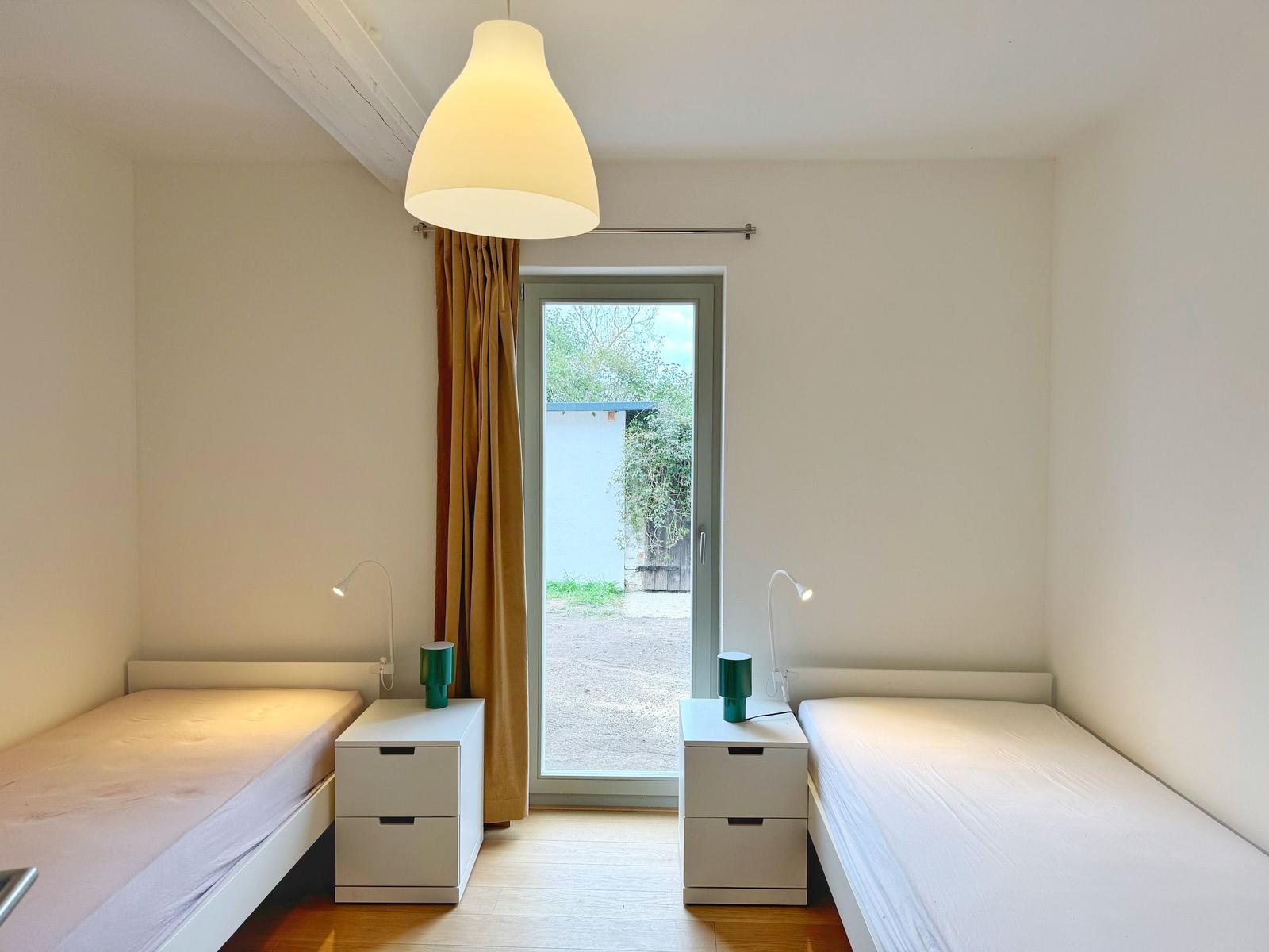 Zwei Einzelbetten mit Nachttischen und Lampen. Tür zum Außenbereich mit Vorhang.