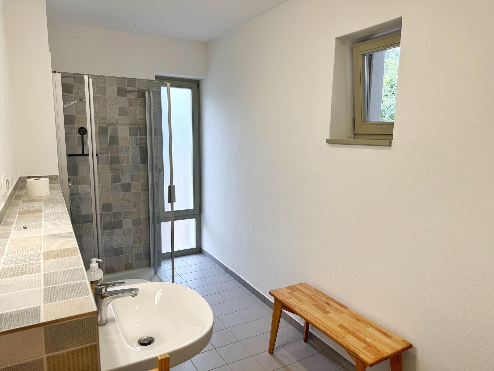 Bad mit Dusche, Waschbecken und Holzbank. Fenster mit Blick nach außen.