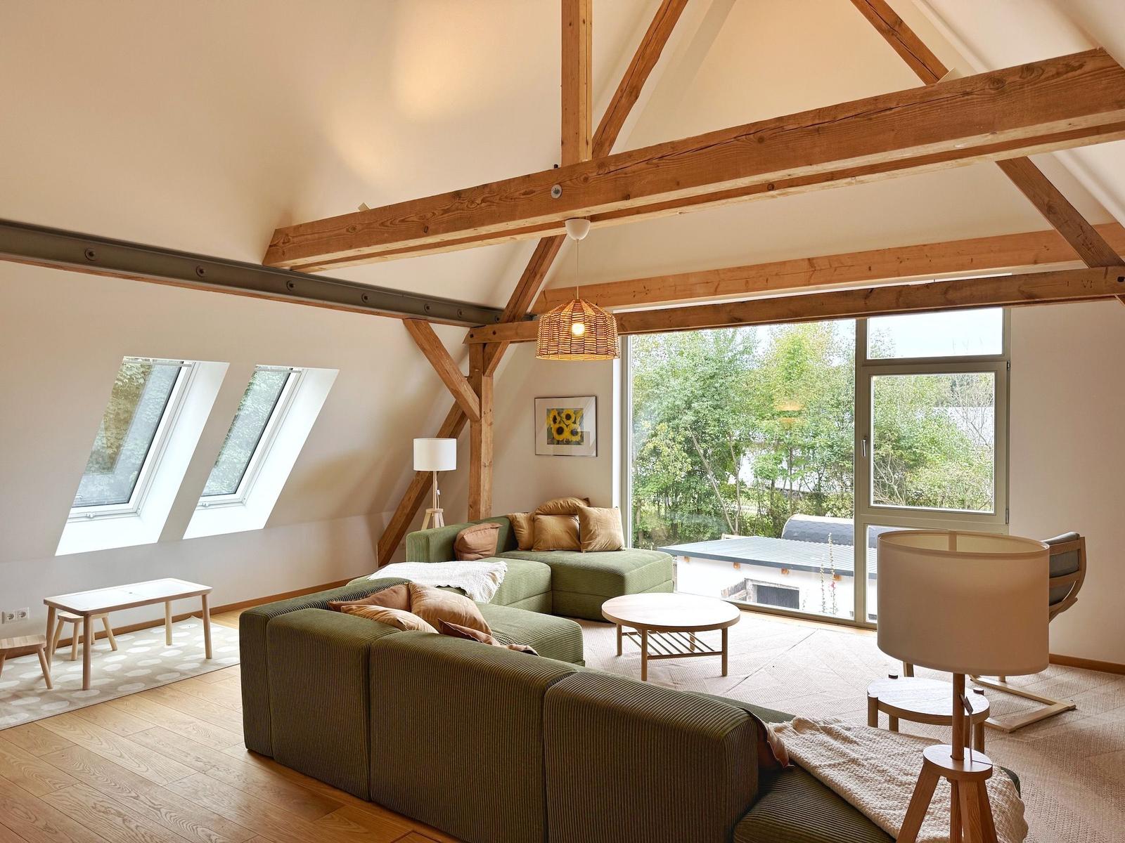 Wohnzimmer mit grünem Sofa, Holzbalken und großen Fenstern zur Außenterrasse.