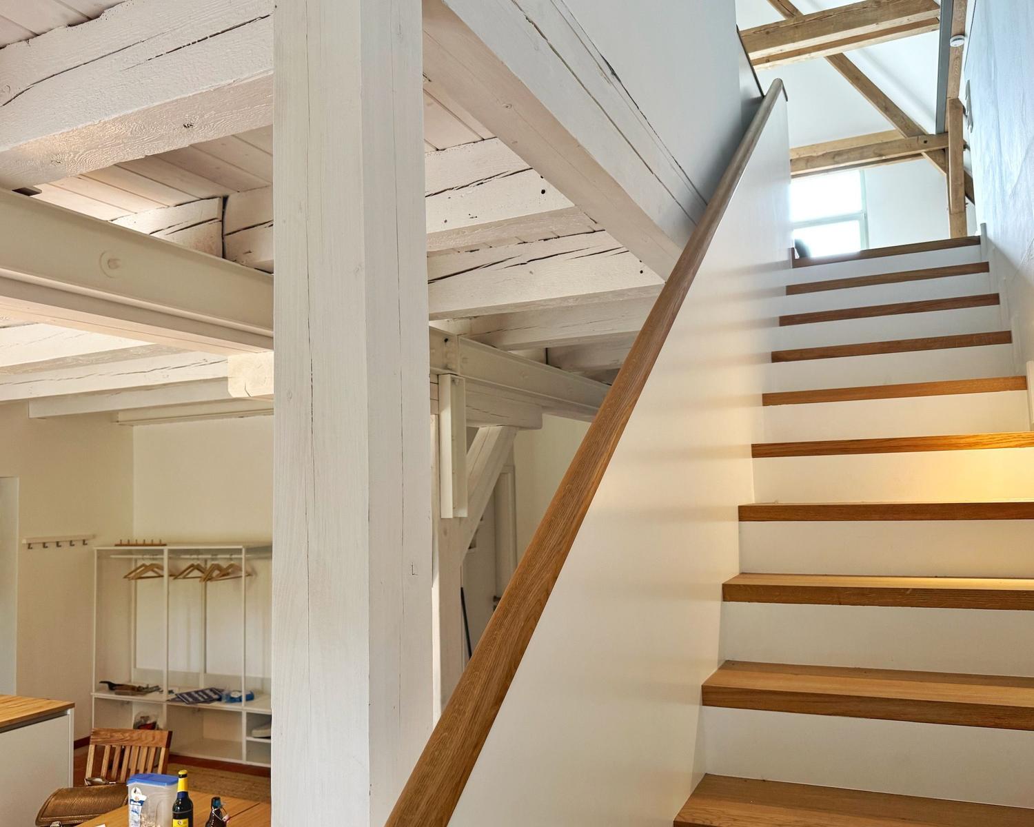 Treppenhaus mit Holzstufen und weiß gestrichenen Balken. Im Hintergrund ist ein Raum mit Kleiderständern.