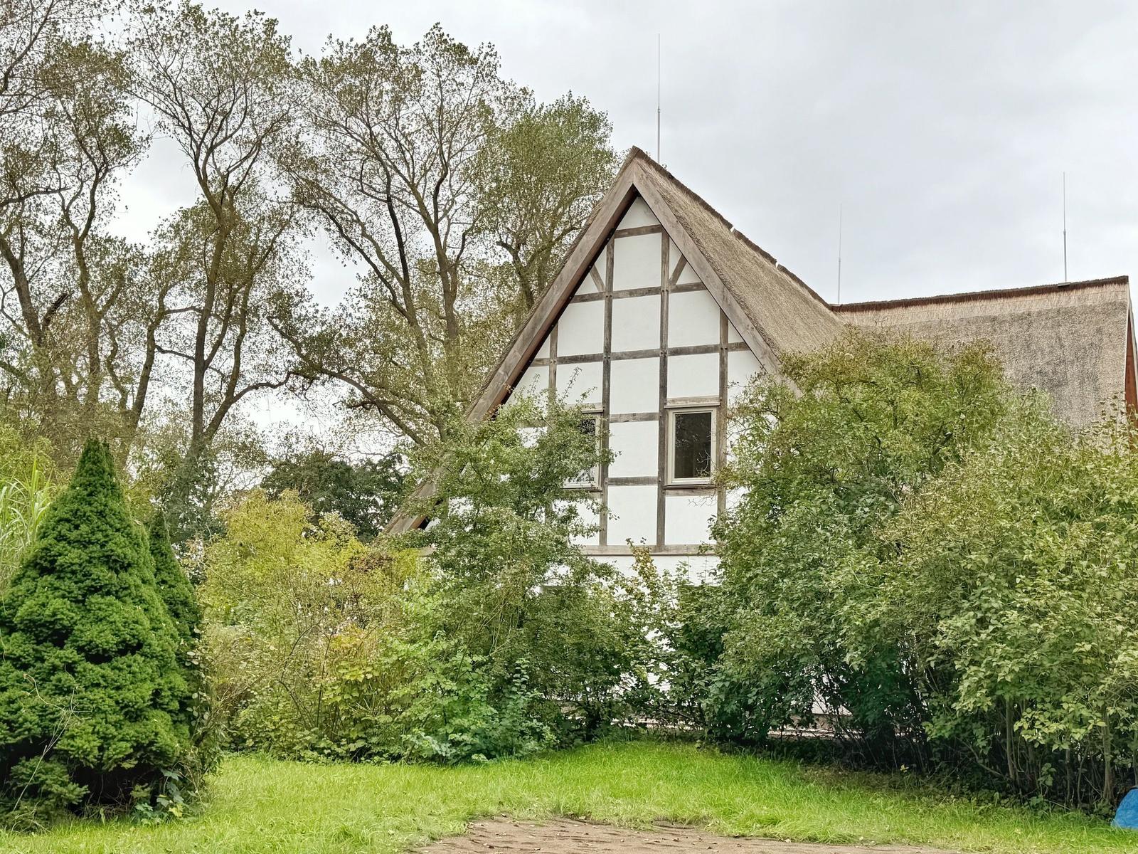 Haus mit Strohdach und Fachwerk, umgeben von Bäumen und Grün.