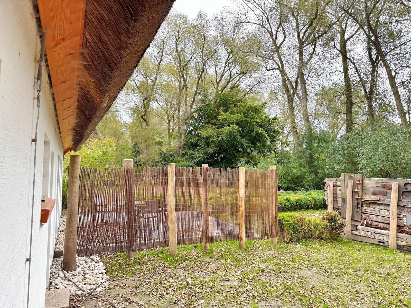 Hinter dem Haus befindet sich ein Garten mit Holzzaun und Tisch. Im Hintergrund sind Bäume zu sehen.