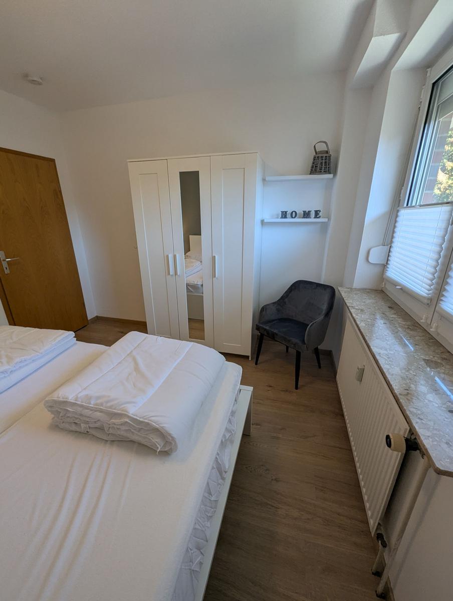 Schlafzimmer mit Bett, Kleiderschrank und Sitzgelegenheit neben Fenster.