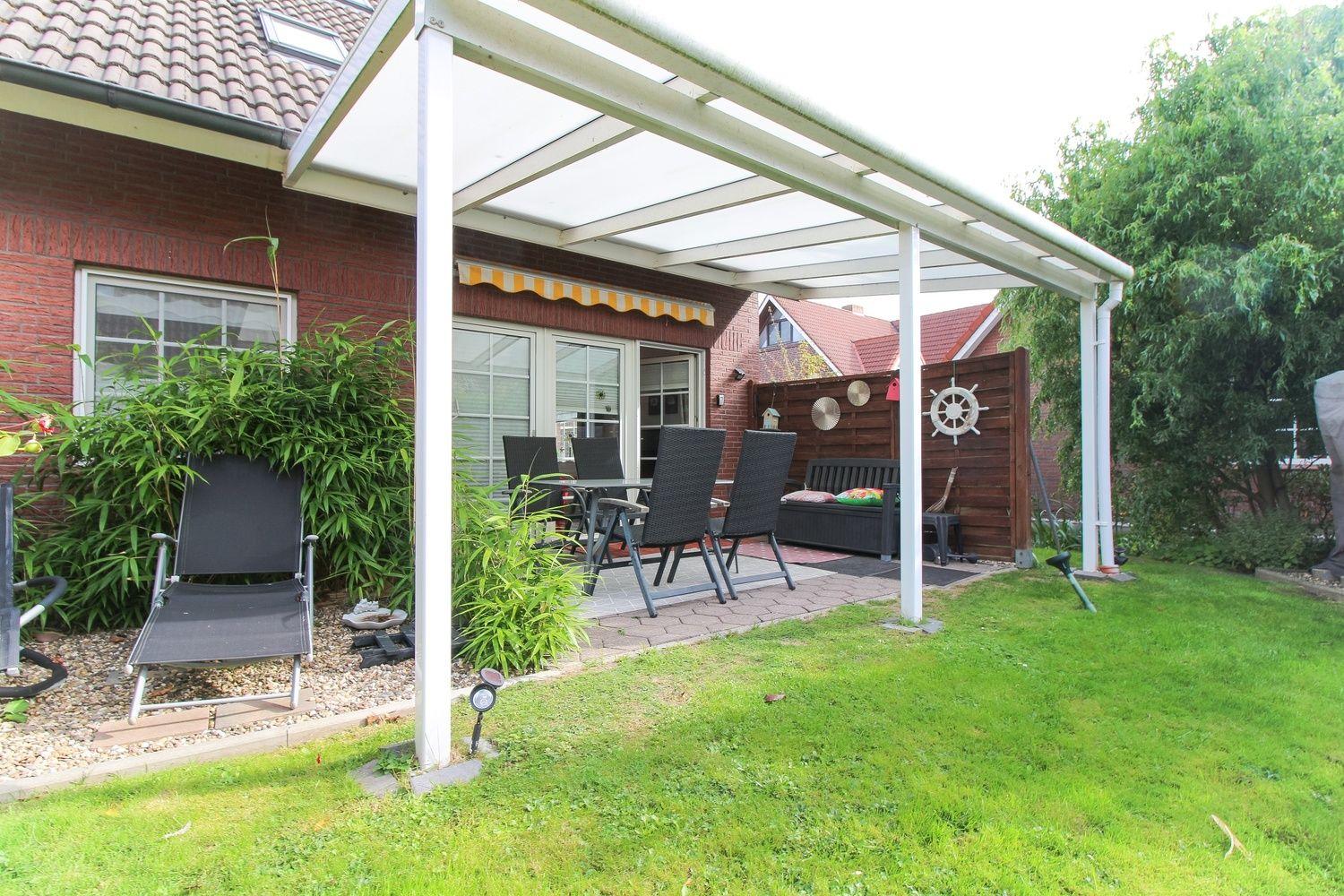 Terrasse mit Sitzgelegenheiten unter einem weißen Dach, umgeben von Grünflächen und einem Holzzaun.