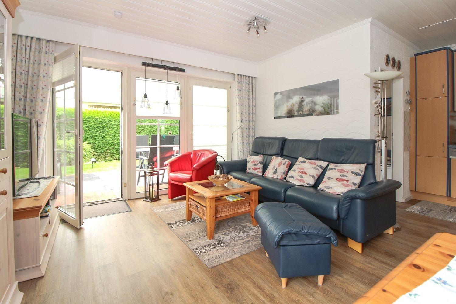 Wohnzimmer mit blauem Sofa, rotem Sessel und Holztisch. Durchgang zur Terrasse.