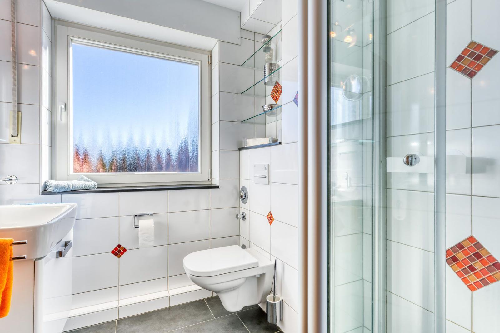 Badezimmer mit Dusche, Toilette und Waschbecken. Fenster mit Blick auf Wald.