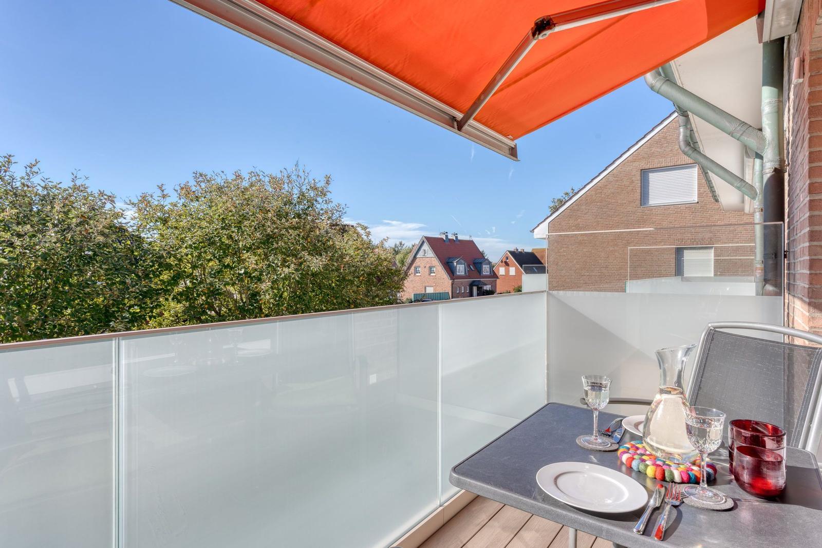 Terrasse mit orangefarbenem Markisenüberdachung und Glastrennwand. Tisch mit Geschirr und Gläsern.