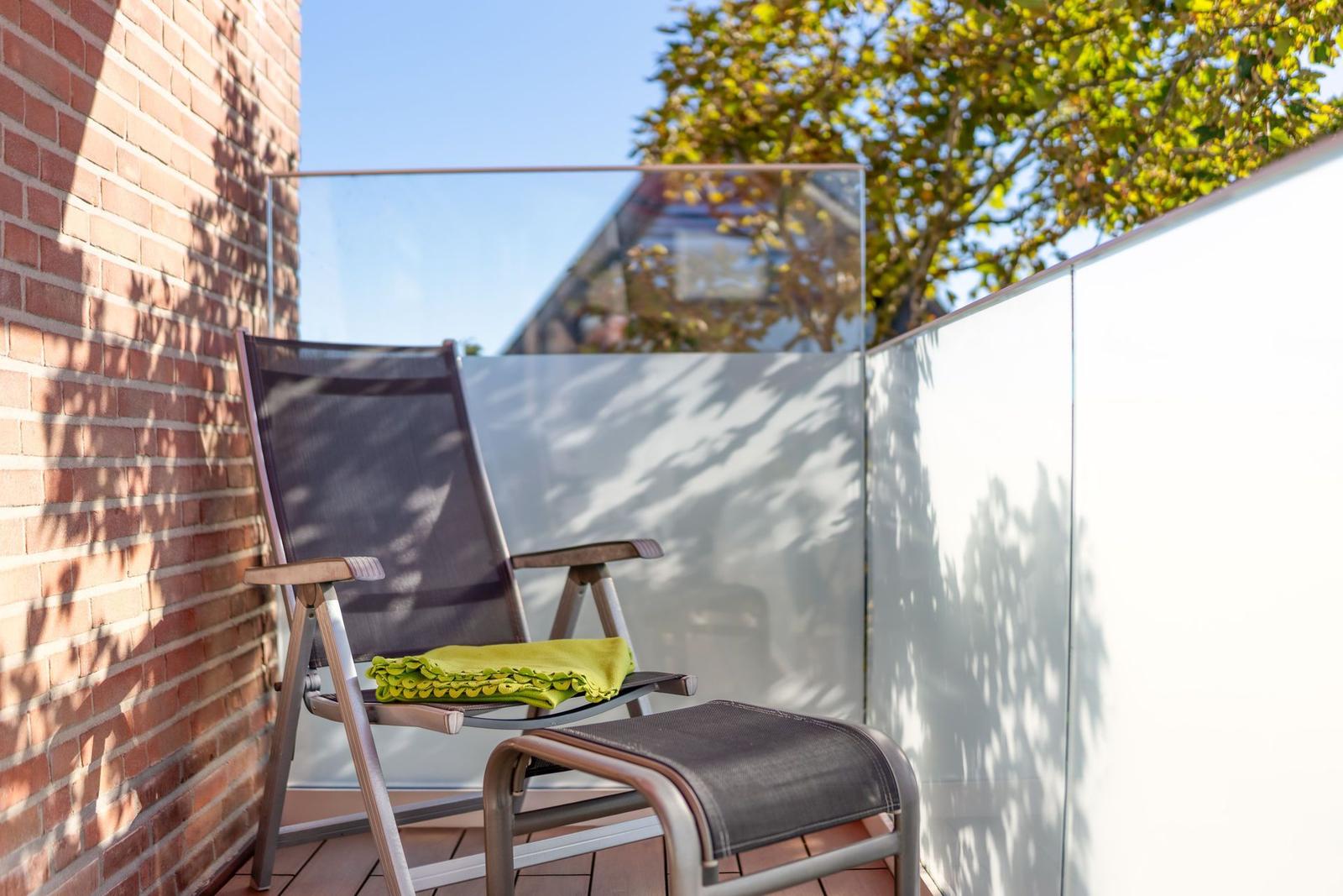 Terrasse mit Liegestuhl und Fußstütze, grüner Decke, Backsteinwand und Glasgeländer.