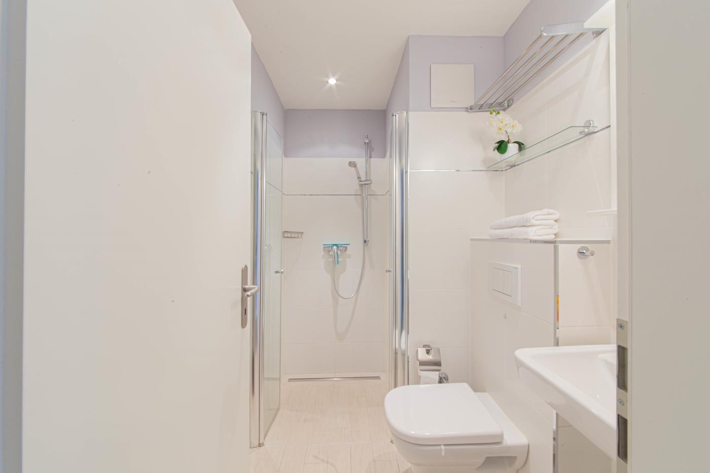 Badezimmer mit Dusche, Toilette und Waschbecken. Duschwand aus Glas. Handtuch und Blume auf Regal.