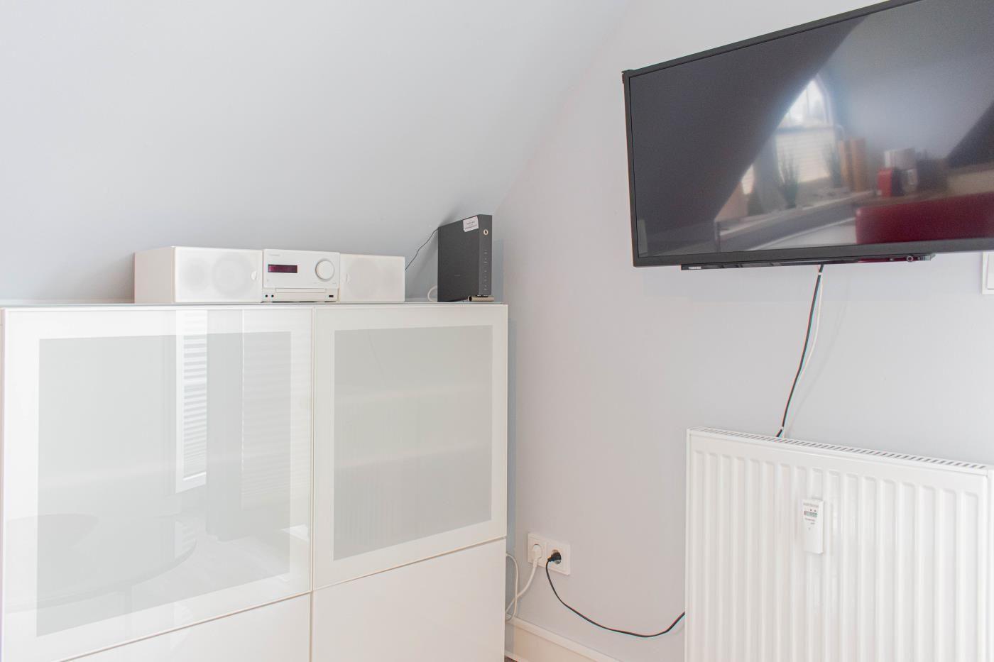 TV an Wand montiert, daneben Weißes Regal mit Audio-Geräten.