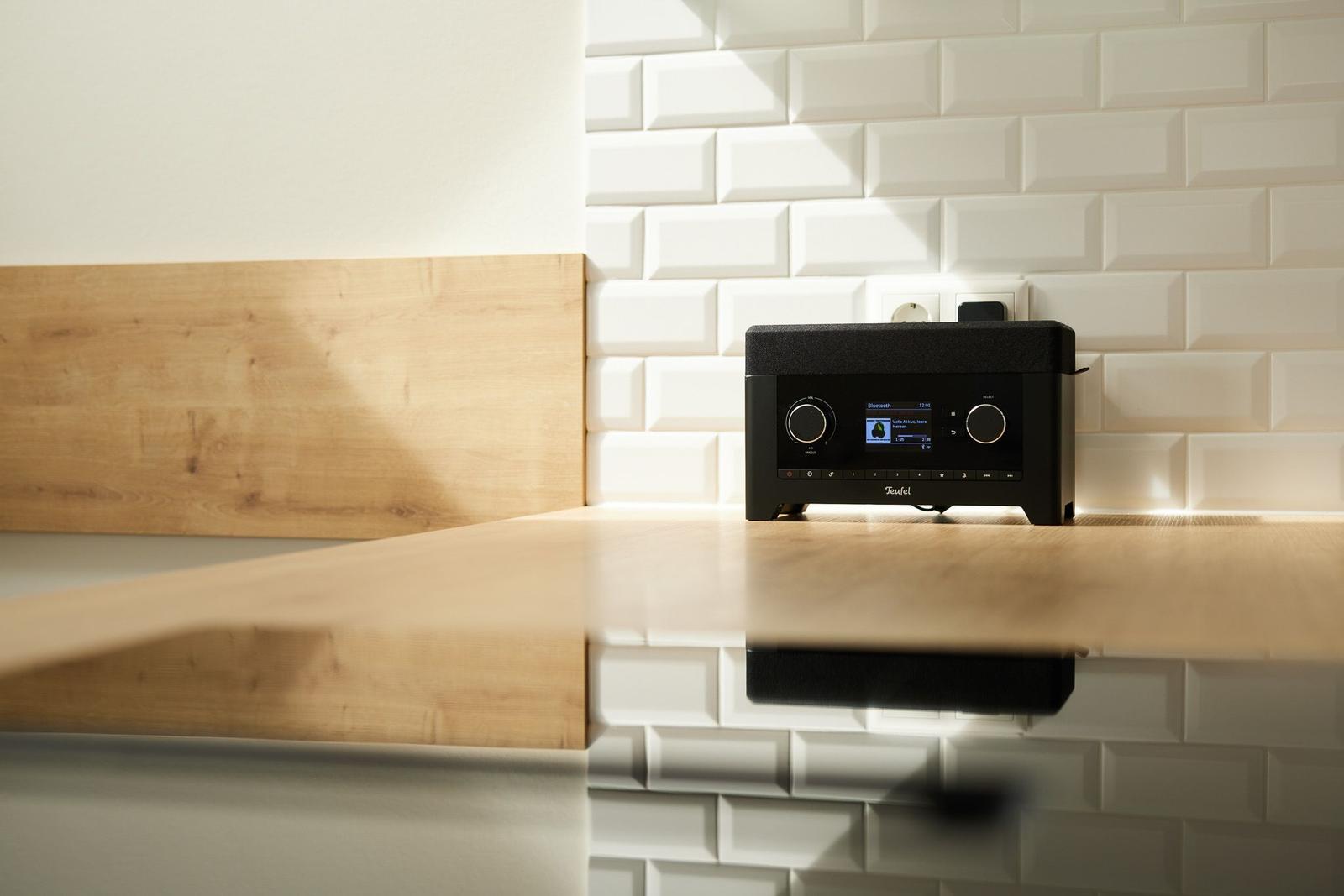 Ein schwarzes Audio-Gerät steht auf einer Holzoberfläche vor einer weißen Fliesenspiegelwand.