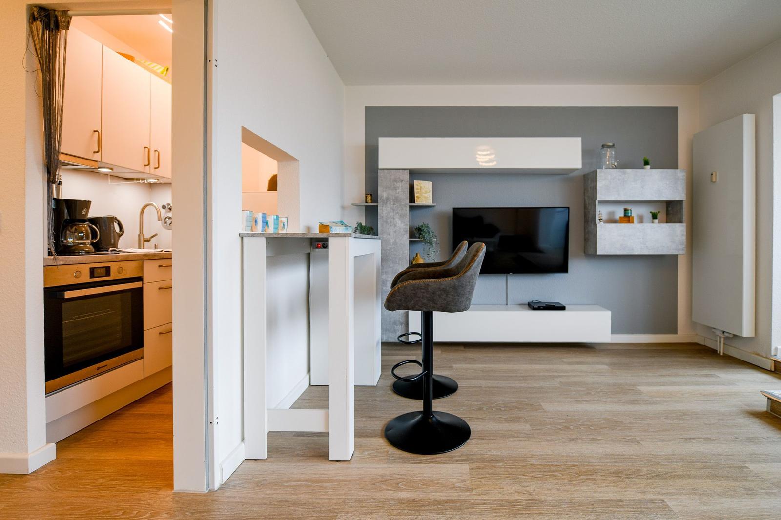 Küche mit Arbeitsplatz und Wohnzimmer mit TV und Barhocker.
