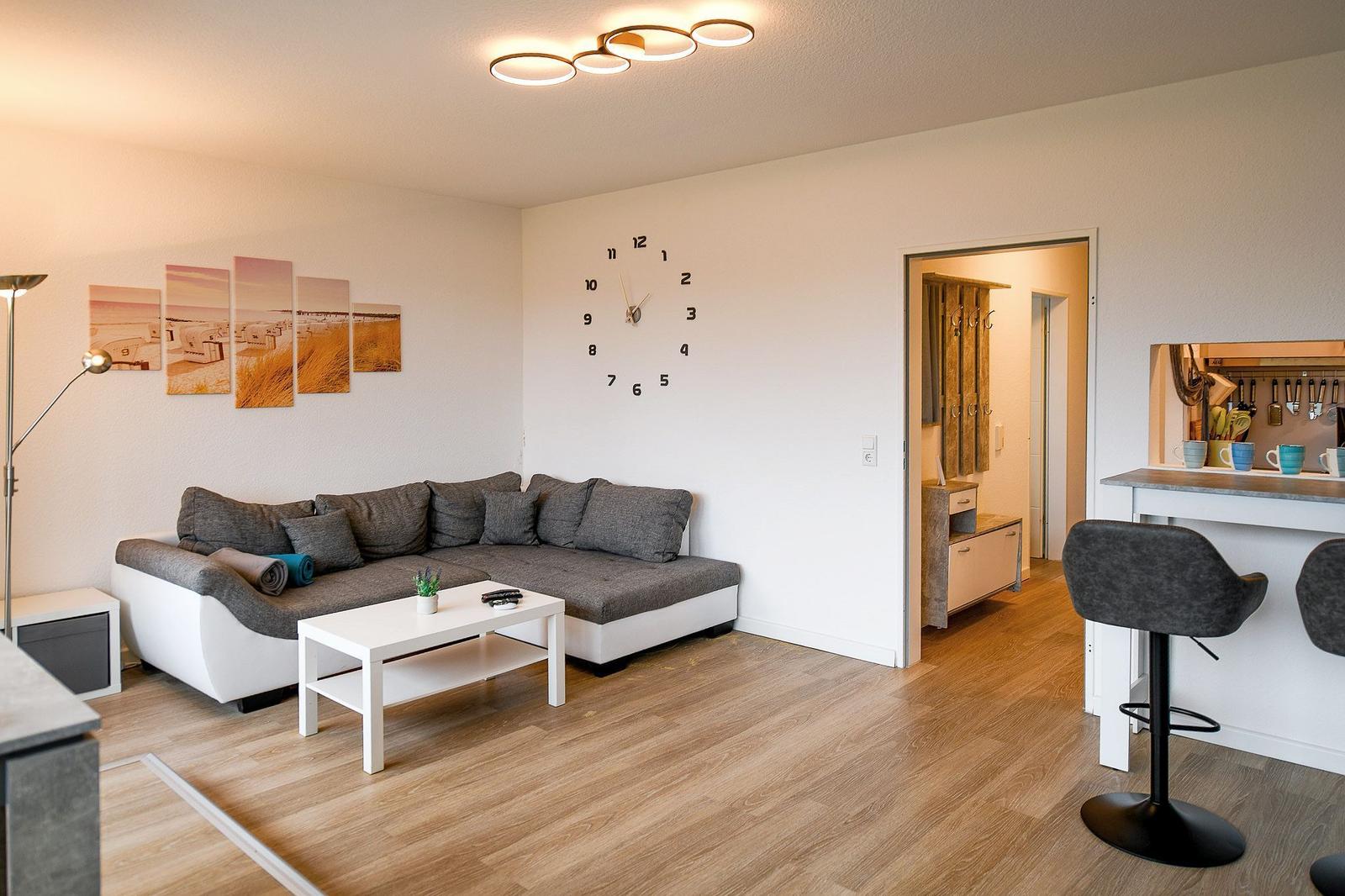 Wohnzimmer mit Sofa, Tisch und Wanduhr. Küche mit Barhocker sichtbar.