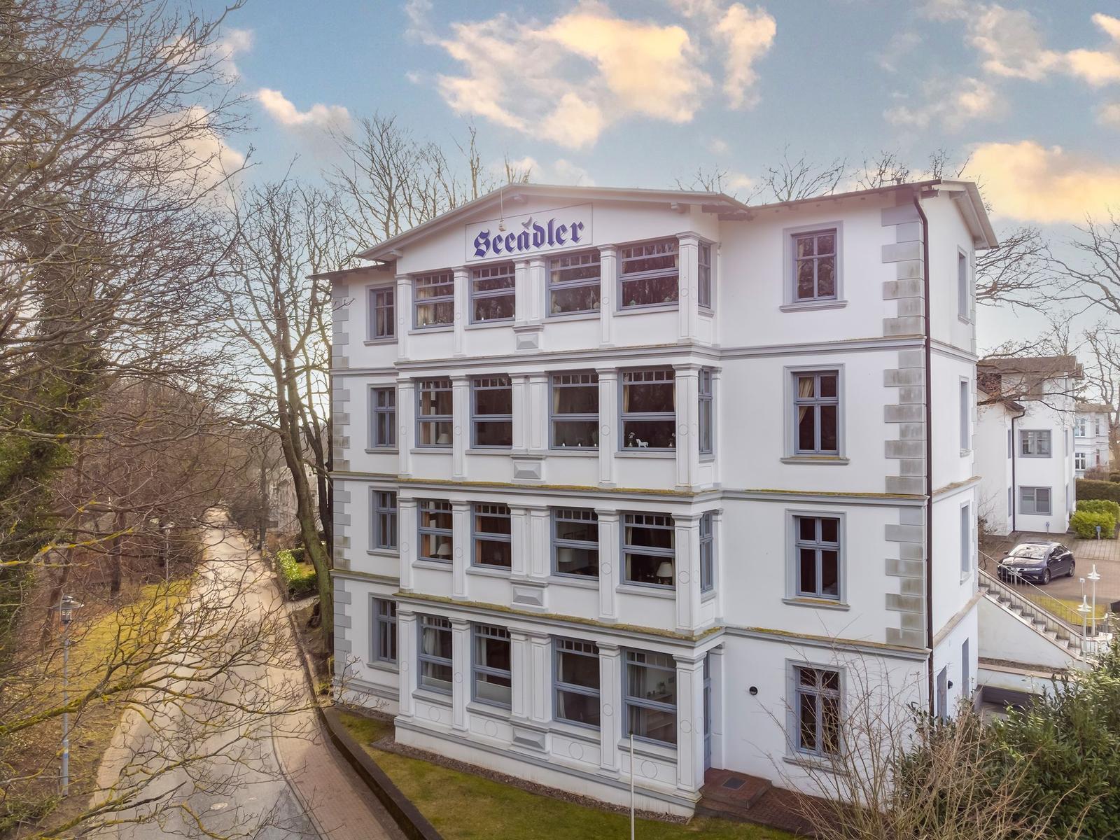Weißes Mehrfamilienhaus mit blauem Schild und Fenstern. Umgeben von Bäumen und Grünflächen.