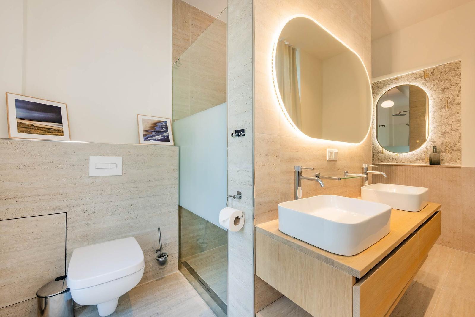 Badezimmer mit Dusche, Toilette und zwei Waschbecken mit beleuchteten Spiegeln.