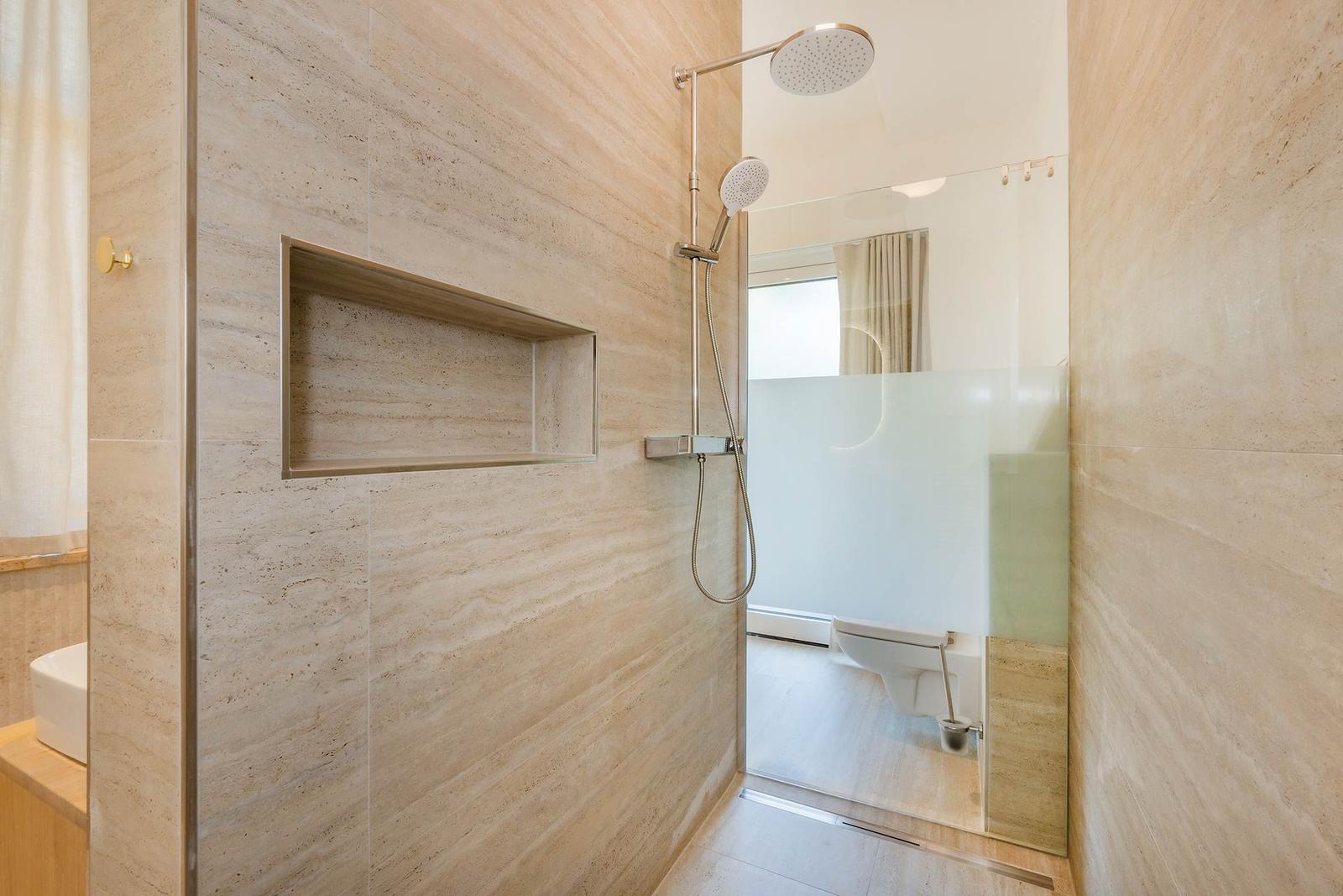 Dusche mit Duschbrause, eingelassene Ablage und Toilette hinter Glaswand.