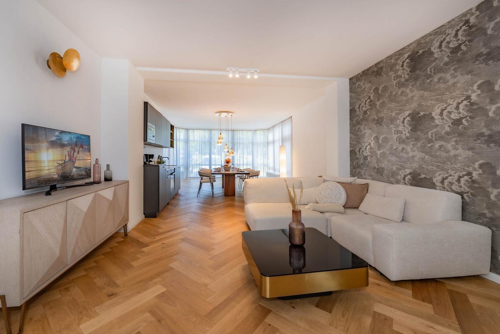 Wohnzimmer mit Sofa, Couchtisch und TV. Küche und Essbereich im Hintergrund.