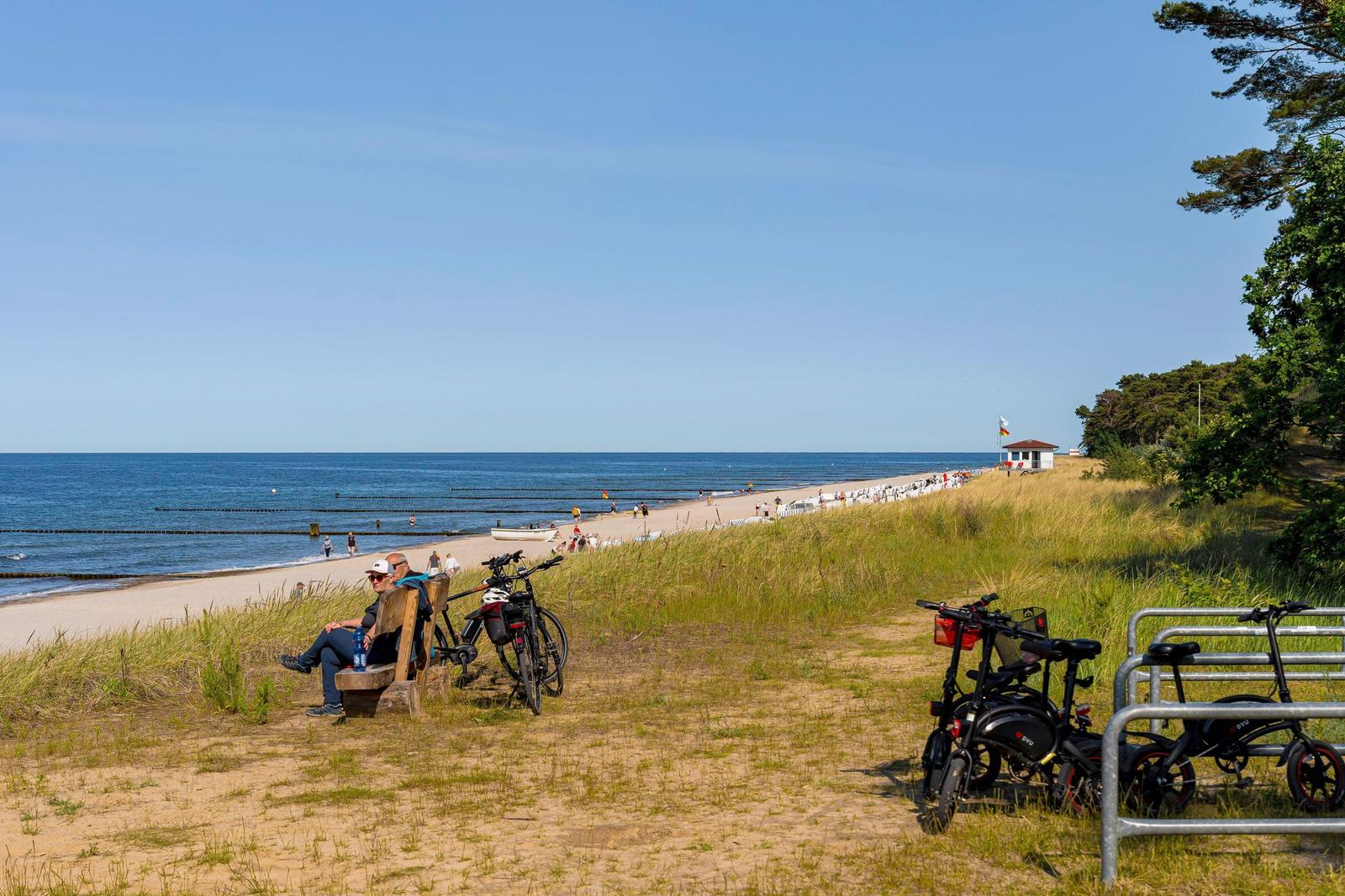 Zwei Personen sitzen auf einer Bank am Strand. Fahrräder stehen daneben.