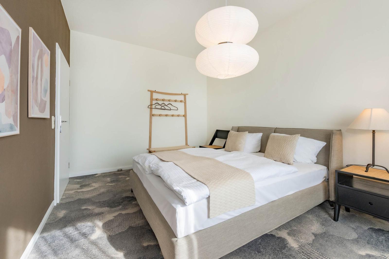 Schlafzimmer mit Bett, Nachttisch und Decke. An der Wand hängen Bilder.