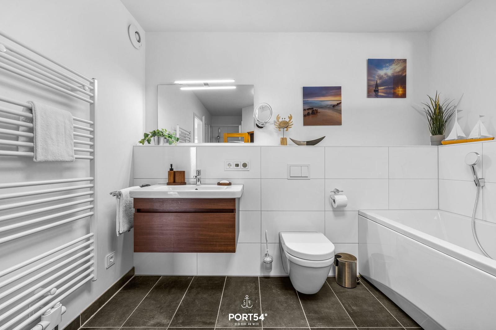 Badezimmer mit Badewanne, Waschbecken und Toilette. Weiße Fliesen und Holzfront.