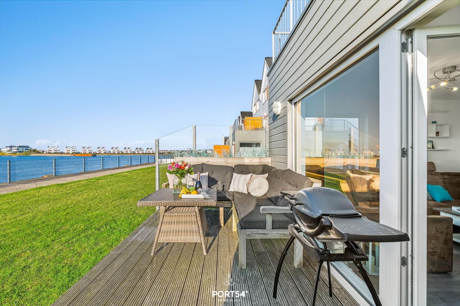Terrasse mit Sofa, Tisch und Grill. Blick auf Wasser und Grünfläche.