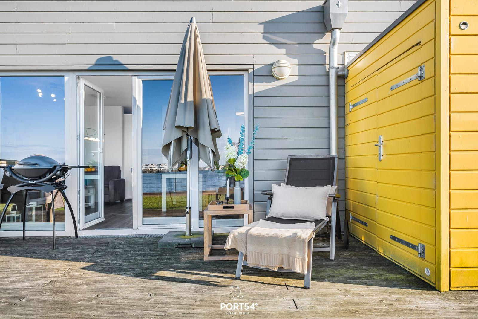 Terrasse mit Liegestuhl, Grill und geschlossenem Sonnenschirm neben gelbem Schuppen.