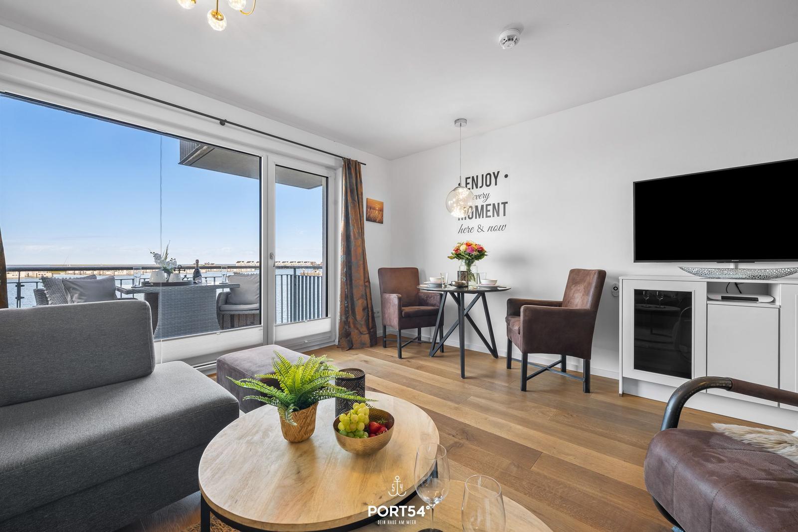 Wohnbereich mit Balkon, TV und Esstisch. Holzboden und große Fenster.