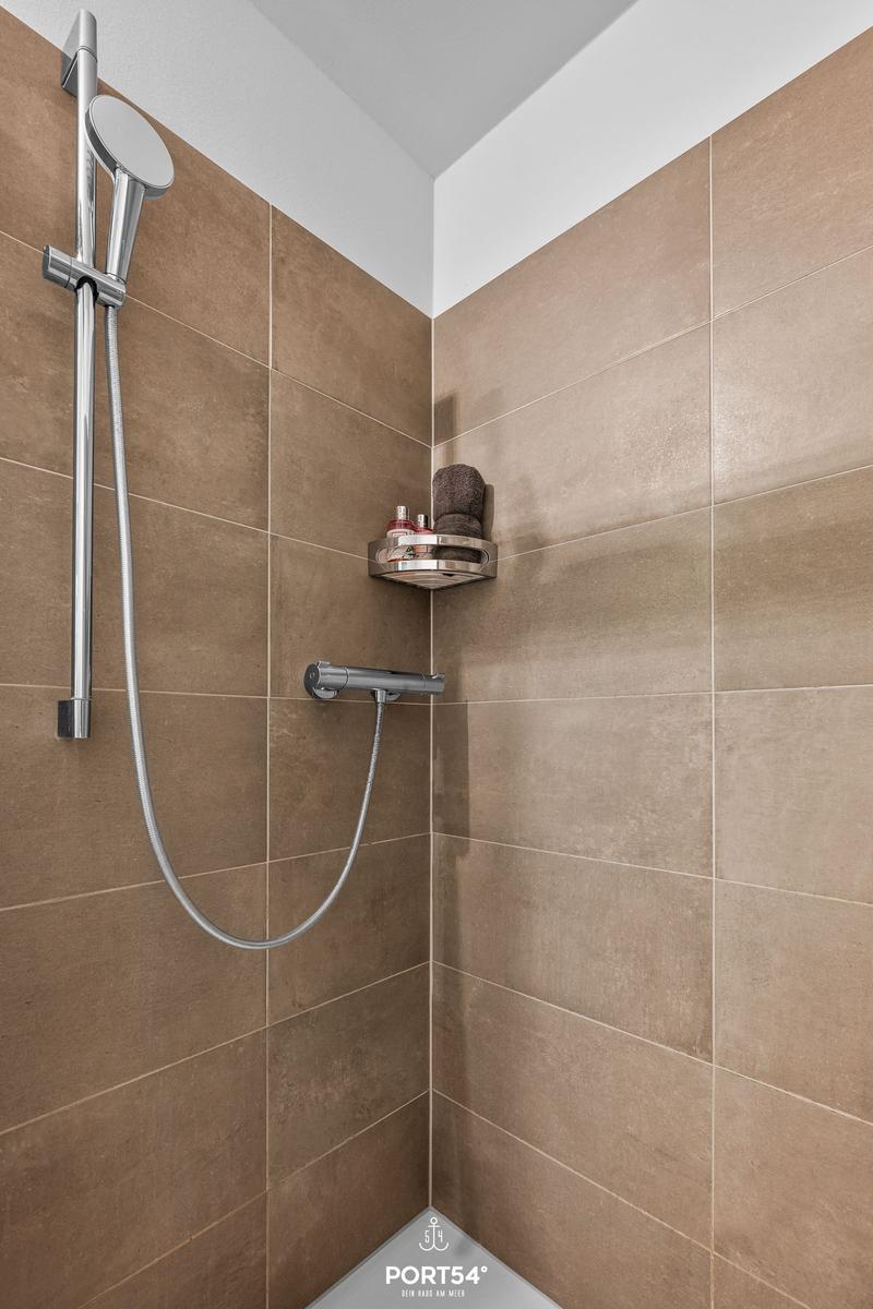 Dusche mit Edelstahlbrause und Handbrause, braune Fliesen, Duschablage mit Handtuch und Flaschen.