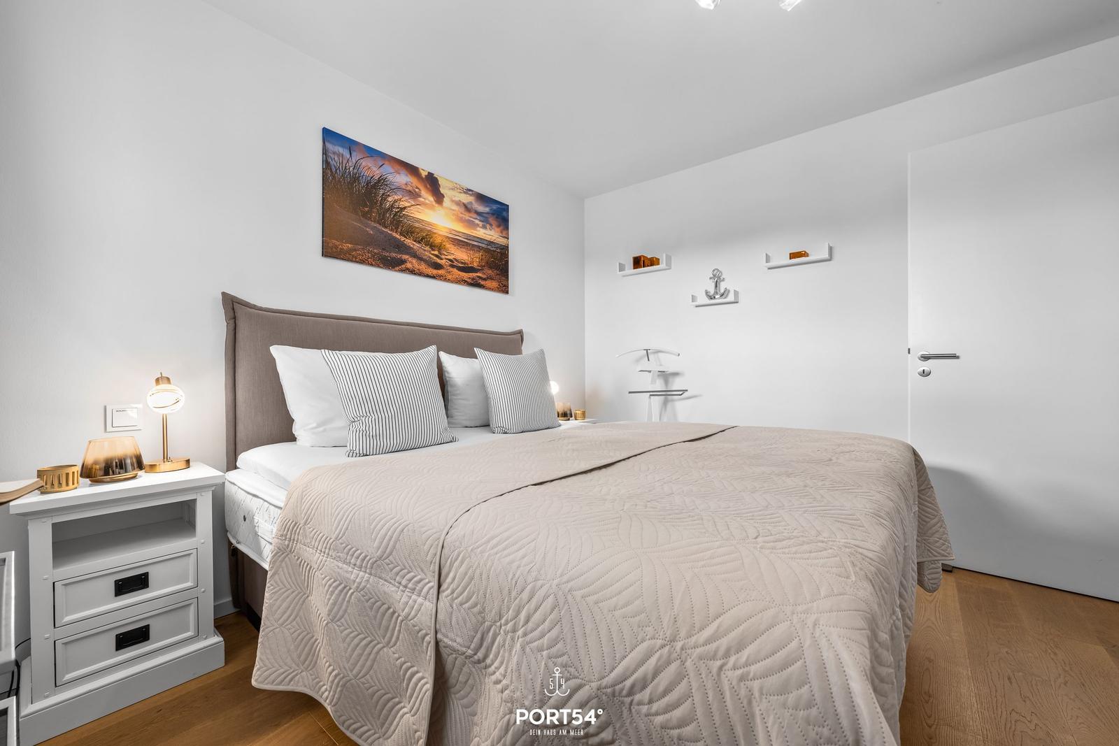 Schlafzimmer mit Bett, Nachttisch und Wandbild. Holzboden und weiße Wände.