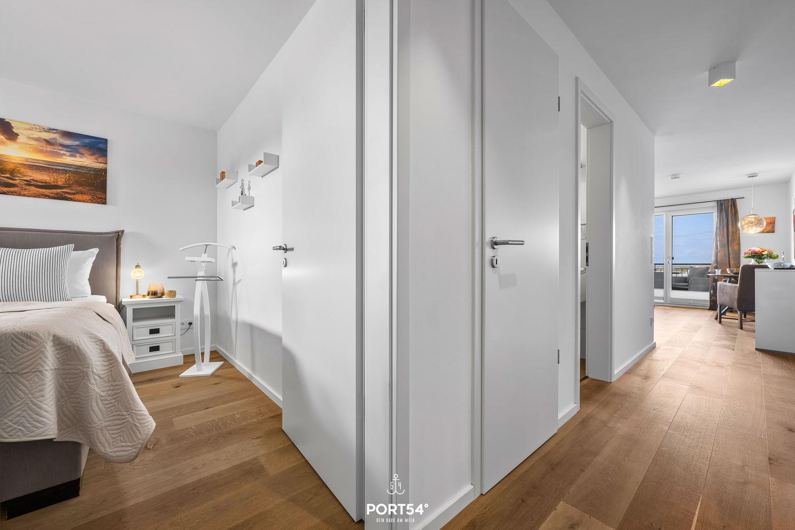 Schlafzimmer mit Bett, Nachttisch und Wandbild. Flur führt zum Wohnbereich mit Balkon.