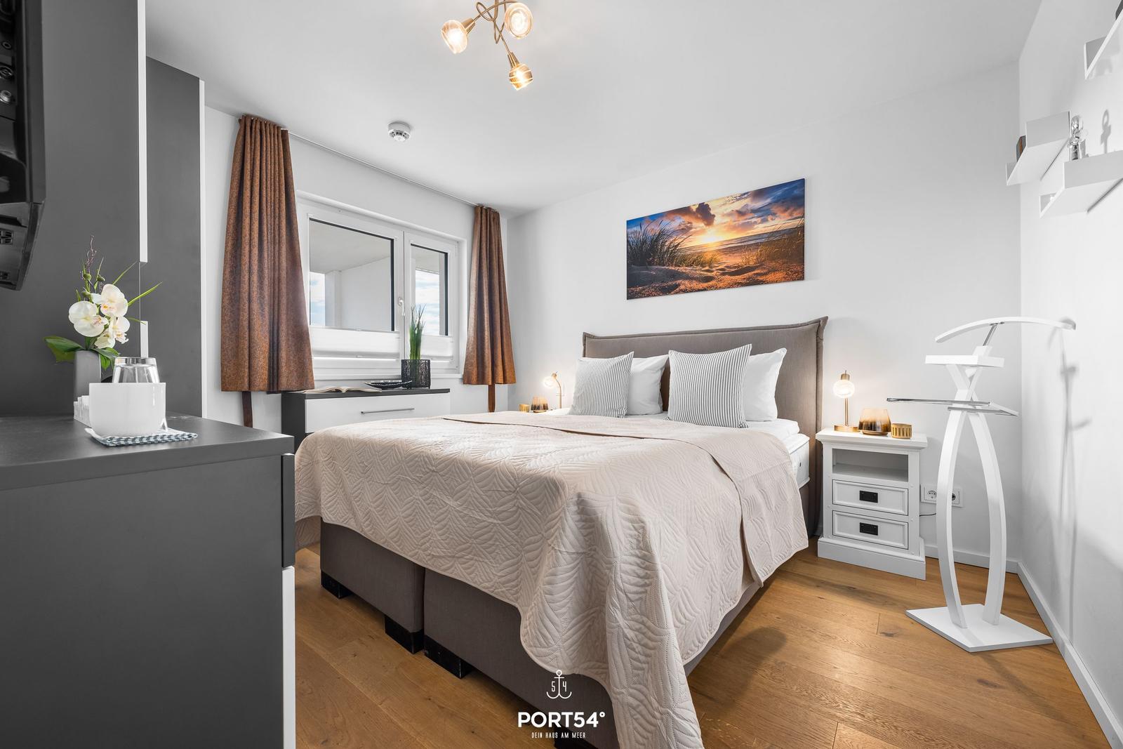 Schlafzimmer mit Bett, Nachttisch und Wandbild. Holzboden und Fenster mit Vorhängen.