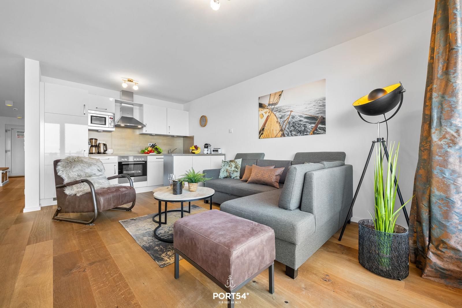 Wohnbereich mit Sofa, Sessel und Küchenzeile. Wandbild und Stehlampe sichtbar.