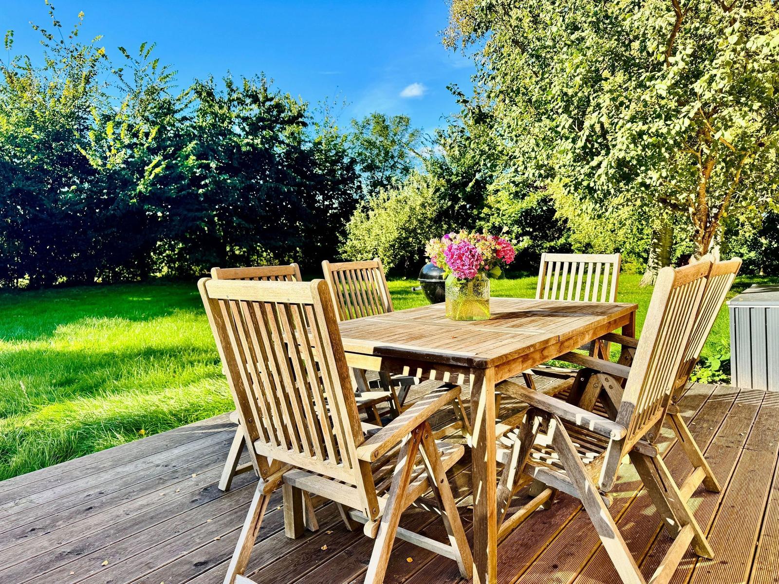 Holztisch mit Stühlen auf Terrasse, Blumen und Grill im Hintergrund.