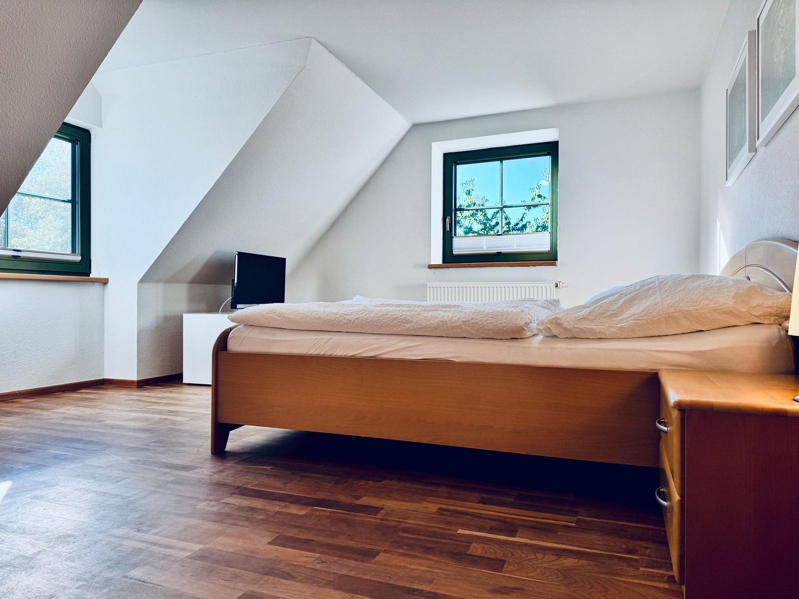 Schlafzimmer mit Dachschrägen, Holzboden und zwei Fenstern mit grünen Rahmen.