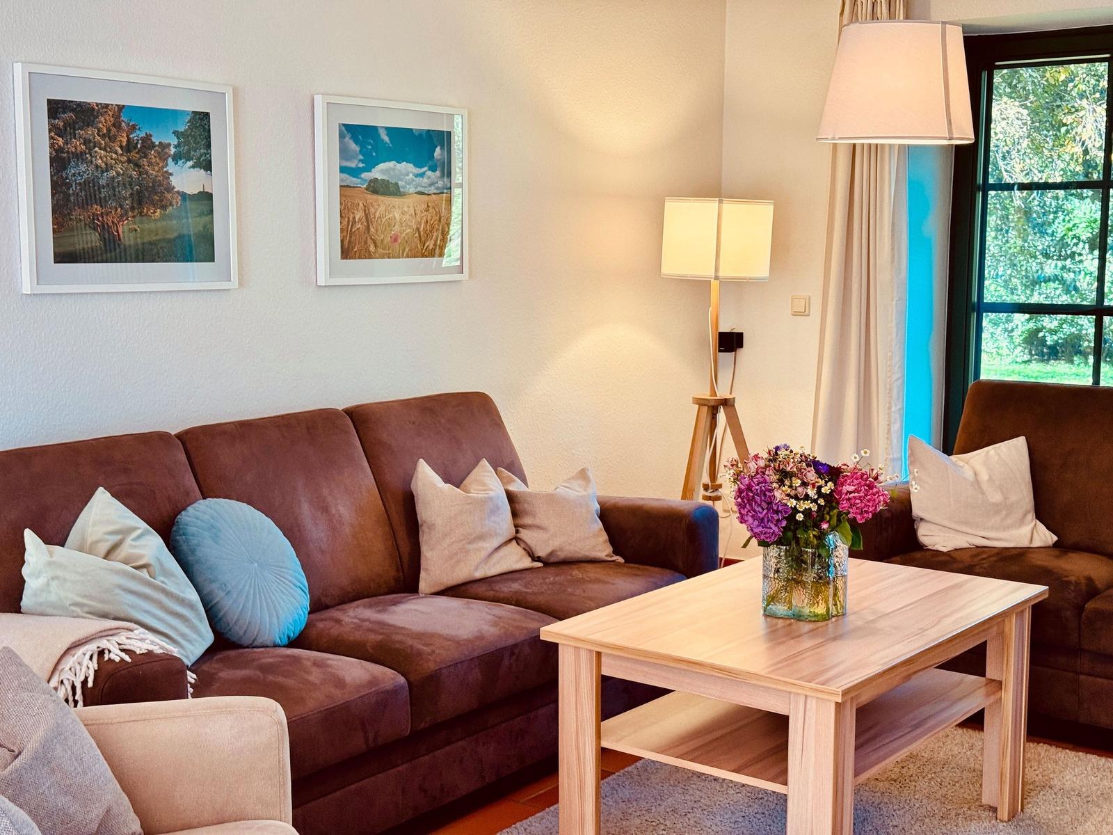 Wohnzimmer mit braunem Sofa, Holztisch und Blumen. An der Wand hängen zwei Bilder.