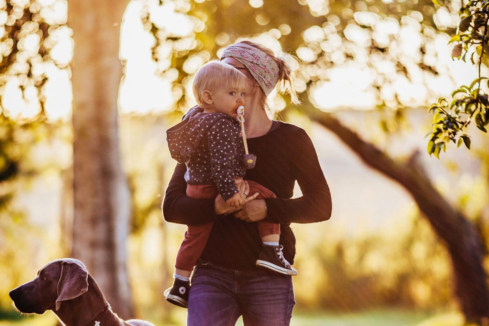 Eine Frau hält ein Kind im Freien. Ein Hund steht daneben. Bäume und Sonnenlicht sind sichtbar.
